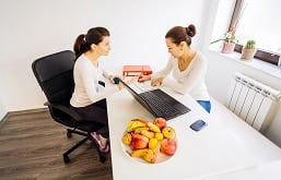 imagem de duas pessoas se conversando em uma mesa onde tem uma cesta de frutas e um notebook.