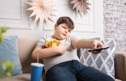 Menino com sobrepeso comendo salgadinho e tomando refrigerante enquanto assiste televisão