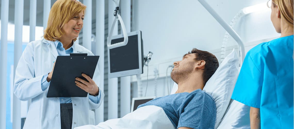 Paciente deitado em leito hospitalar enquanto médica conversa com ele acompanhado de enfermeira.