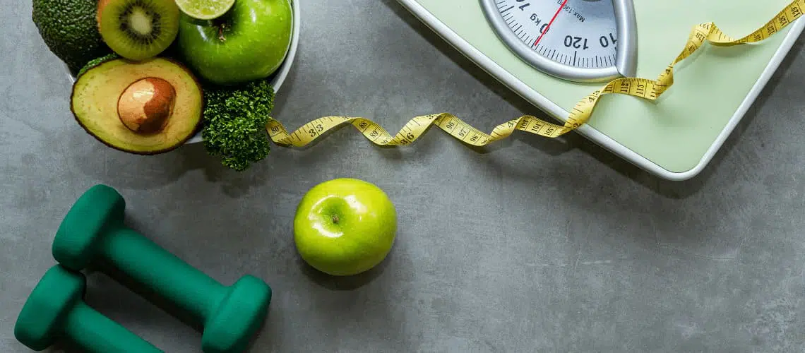 Foto com vista aérea de uma balança, fita métrica, abacates, maçã verde e pesos para exercícios.