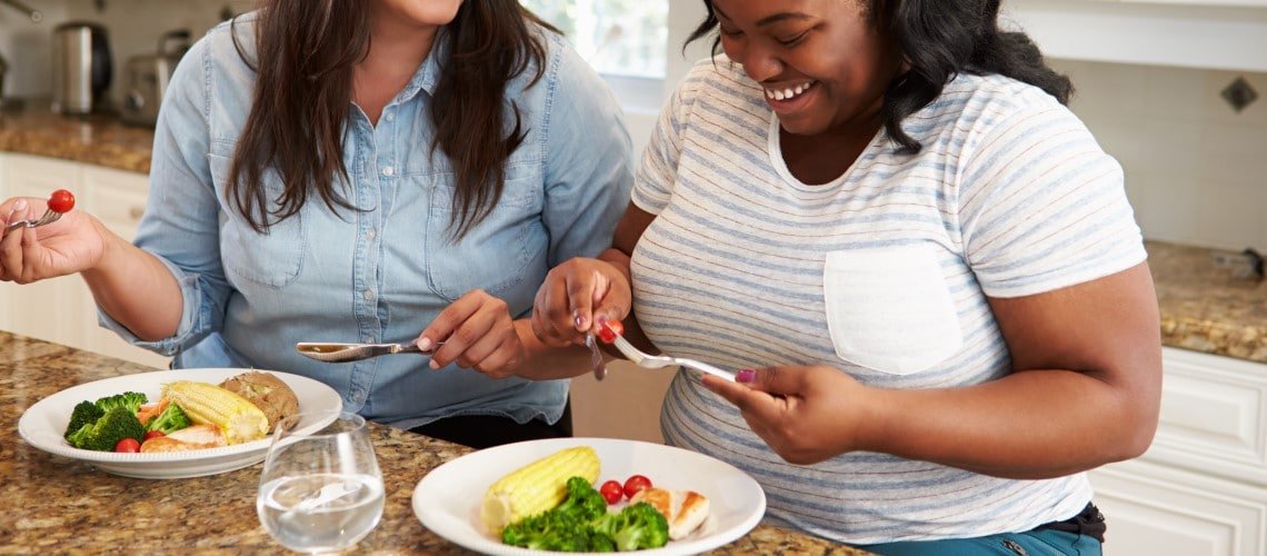 duas mulheres segurando talheres, balcão com dois pratos contendo milho, brócolis, tomate cereja e frango