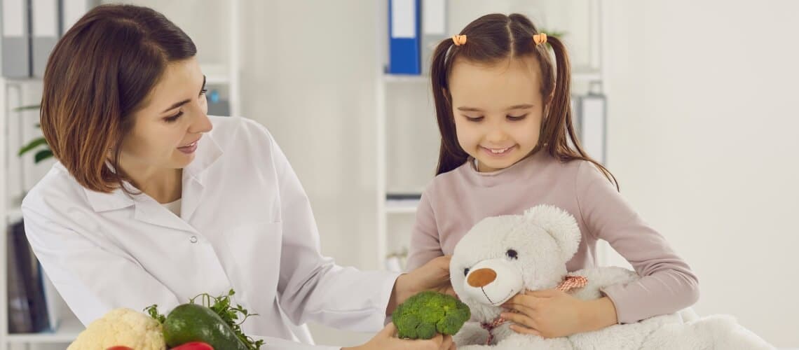menina segurando urso de pelúcia e mulher com jaleco mostrando brócolis para menina