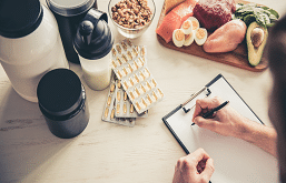 imagem de uma pessoa escrevendo em uma prancheta com frutas e uma garrafa de café do lado.