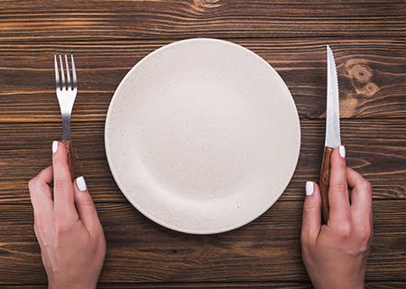 Mãos segurando talheres ao lado de prato vazio sobre a mesa