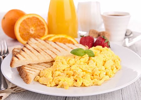 Pão com ovos mexidos e fatias de pão e, ao fundo, copos com suco de laranja e leite; uma mesa de café da manhã