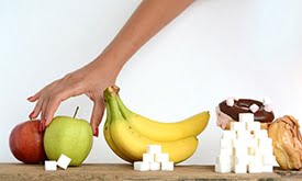 Mesa com pera, banana e rosquinhas e cubinhos de açúcar em frente cada um deles, indicando a quantidade de açúcar que contém. Mão feminina pegando a pêra.