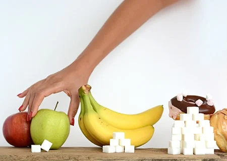 Mesa com pera, banana e rosquinhas e cubinhos de açúcar em frente cada um deles, indicando a quantidade de açúcar que contém. Mão feminina pegando a pêra.