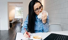 Mulher jovem escrevendo em caderno com uma mão e, com outra, comendo um lanche