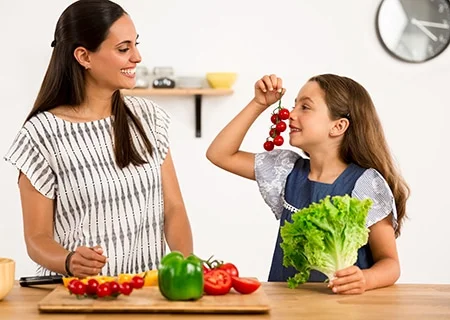 Mulher e garotinha atrás de bancada repleta de verduras na cozinha, se olhando e sorrindo. Garota segurando um pé de alface e tomates com as mãos.