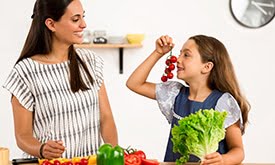 Mulher e garotinha atrás de bancada repleta de verduras na cozinha, se olhando e sorrindo. Garota segurando um pé de alface e tomates com as mãos.