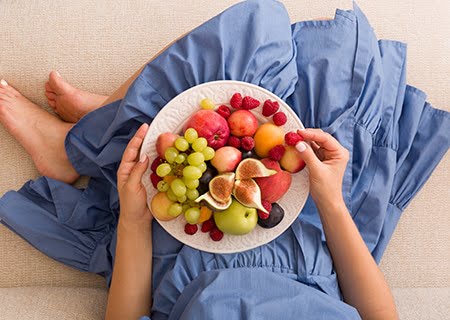 Mulher sentada, com um prato repleto de diferentes frutas no seu colo