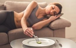 Mulher deitada no sofá com expressão triste dando garfada em um prato que contém apenas um pequeno pedaço de legume