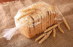 Pão de fôrma fatiado sobre mesa, em embalagem de plástico sobre saco de juta e ramos de trigo ao lado