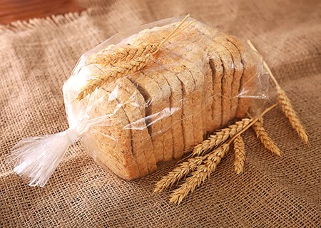 Pão de fôrma fatiado sobre mesa, em embalagem de plástico sobre saco de juta e ramos de trigo ao lado