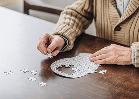Mão de idosa montando quebra-cabeça sobre mesa. O desenho do jogo é uma face, e faltam as peças da área referente ao cérebro.
