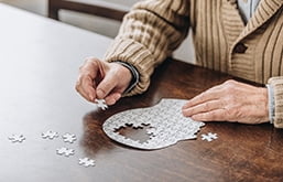 Mão de idosa montando quebra-cabeça sobre mesa. O desenho do jogo é uma face, e faltam as peças da área referente ao cérebro.