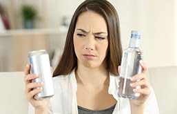 Mulher segurando uma bebida dietética em uma mão e, na outra, uma garrafa de água, com expressão de dúvida sobre o que tomar