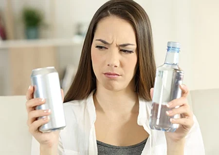 Mulher segurando uma bebida dietética em uma mão e, na outra, uma garrafa de água, com expressão de dúvida sobre o que tomar