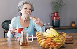 Mulher idosa sentada sorrindo tomando café da manhã em mesa com café e cesta de frutas