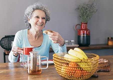 Mulher idosa sentada sorrindo tomando café da manhã em mesa com café e cesta de frutas