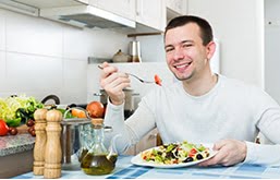 Homem sorrindo sentado à mesa comendo prato cheio de legumes, que faz parte de uma alimentação adequada para prevenir e tratar o câncer de próstata.