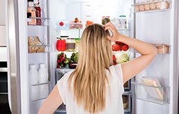 Mulher de costas, olhando para uma geladeira aberta, com a mão na cabeça, pensativa.