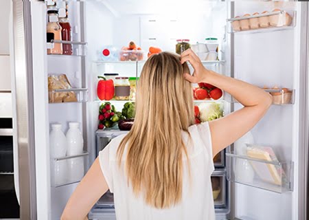 Mulher de costas, olhando para uma geladeira aberta, com a mão na cabeça, pensativa.