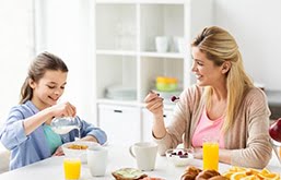 Mãe e filha sentadas à mesa tomando café da manhã e sorrindo, Na mesa tem sucos, leite e pães