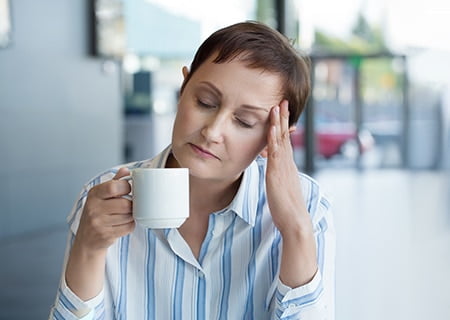 Mulher sentada segura uma xícara de café em uma mão, enquanto apoia a outra mão sobre a testa. De olhos fechados, ela passa a impressão de cansaço e sensação de dor de cabeça