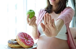 Mulher grávida segurando uma maçã verde e fazendo sinal de negativo com a outra mão para prato com donuts que está ao lado dela