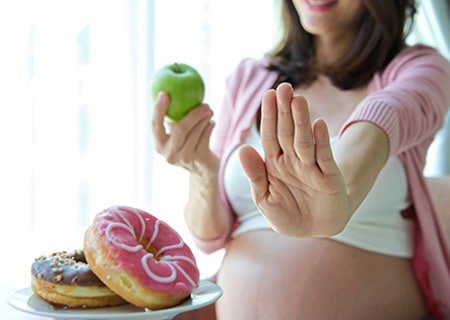 Mulher grávida segurando uma maçã verde e fazendo sinal de negativo com a outra mão para prato com donuts que está ao lado dela