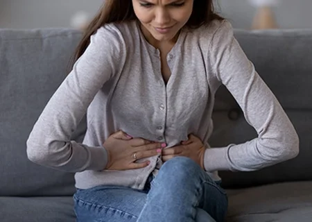 Mulher sentada com as mãos na barriga, contorcida, demonstrando dor. Entre as medidas para aliviar desconfortos abdominais, estão os alimentos com lactobacilos para gastrite