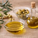Mesa com azeite em garrafa e pote de vidro e outro pote com azeitonas, além de folhas de oliveira espalhadas