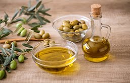 Mesa com azeite em garrafa e pote de vidro e outro pote com azeitonas, além de folhas de oliveira espalhadas