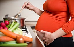 Perfil de mulher grávida cozinhando. Com uma mão, ela mexe uma colher em uma panela no fogão e, com a outra, acaricia a barriga. Ela veste uma blusa laranja.