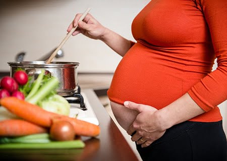 Perfil de mulher grávida cozinhando. Com uma mão, ela mexe uma colher em uma panela no fogão e, com a outra, acaricia a barriga. Ela veste uma blusa laranja.