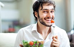 Homem sorri segurando um garfo com um tomate cereja espetado. À sua frente, um pote de vidro com salada de folhas e tomates