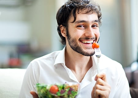 Homem sorri segurando um garfo com um tomate cereja espetado. À sua frente, um pote de vidro com salada de folhas e tomates