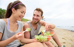 Uma mulher e um homem sentados na praia, com roupas de ginástica, sorrindo e comendo salada