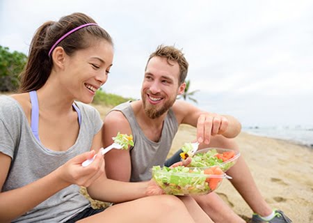 Uma mulher e um homem sentados na praia, com roupas de ginástica, sorrindo e comendo salada
