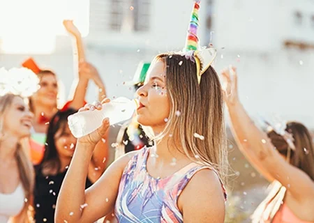 Menina no meio de festa de Carnaval, com uma tiara de unicórnio, bebendo água em garrafa