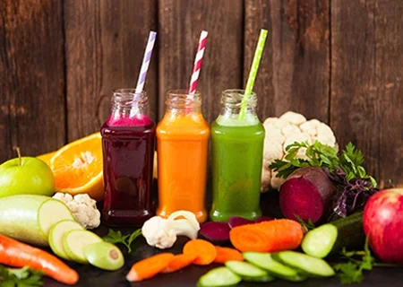 Sucos em garrafas de vidro sobre mesa com vários vegetais, como penino, cenoura e laranja fatiados