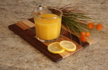 Suco de laranja sobre tábua de madeira ao lado de folhas e fatias de laranja. Imagem de Mira Cosic por Pixabay.