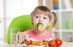 Menina sentada com um prato de verduras à frente. Ela segura um brócolis com o garfo e o leva à boca.