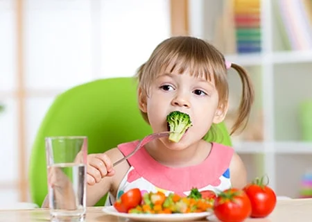 Menina sentada com um prato de verduras à frente. Ela segura um brócolis com o garfo e o leva à boca.