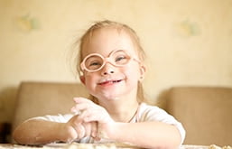 Garotinha com síndrome de Down sorri. Ela usa óculos de armação laranja e redonda e tem as mãos sujas de farinha
