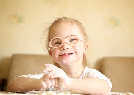 Garotinha com síndrome de Down sorri. Ela usa óculos de armação laranja e redonda e tem as mãos sujas de farinha