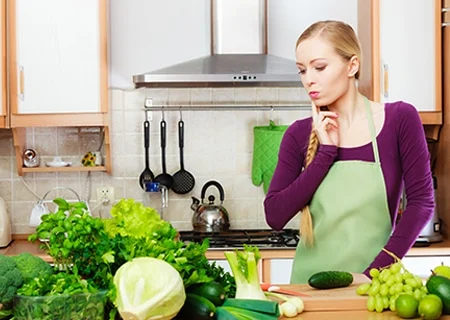 Mulher atrás de bancada na cozinha cheia de verduras com a mão no queixo pensativa indicando dúvida. Ela veste uma blosa roxa de mangas compridas, um avental verde, é loira e tem uma trança lateral.