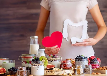 Silhueta de mulher segurando um intestino de papel com uma mão e, com outra, um coração de papel. Ela está atrás de uma mesa repleta de diversos alimentos, como frutas e leite em jarras de vidro.