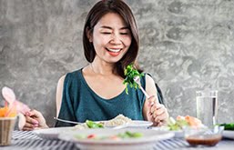 Mulher sentada à mesa segurando um garfo com folhas verdes de salada espetadas.